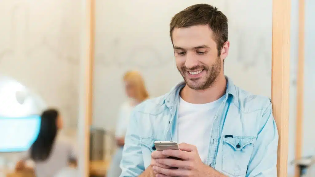 A lightly bearded man smiling while texting on his phone.