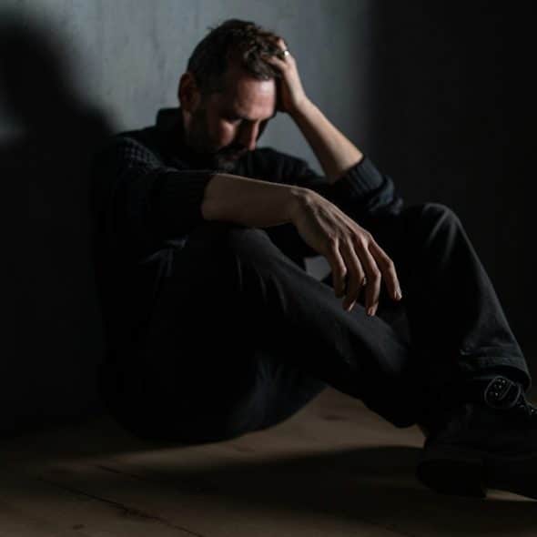 A distressed man sits on the floor in a dark room with his head in his hand.