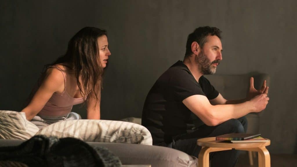 A woman looking at a man sitting beside a bed in a dimly lit room.
