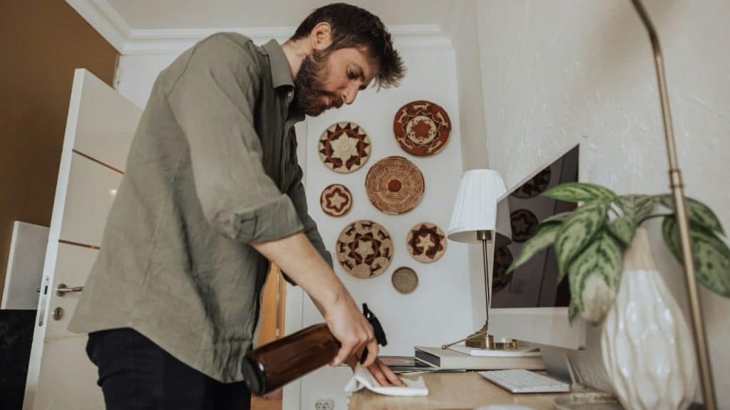 A bearded man in a button-down shirt is cleaning and wiping down a desk.