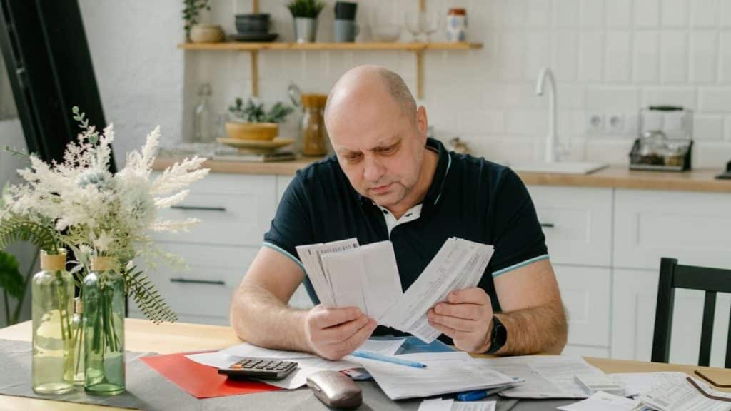 A focused, balding man in a dark polo shirt is sitting at a kitchen table, looking at bills and papers.
