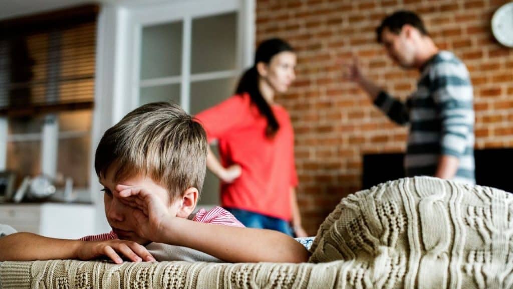 A sad child lying on a sofa while parents argue in the blurred background.