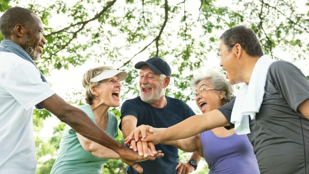 A joyful group of diverse, older friends is laughing and stacking their hands together outdoors.