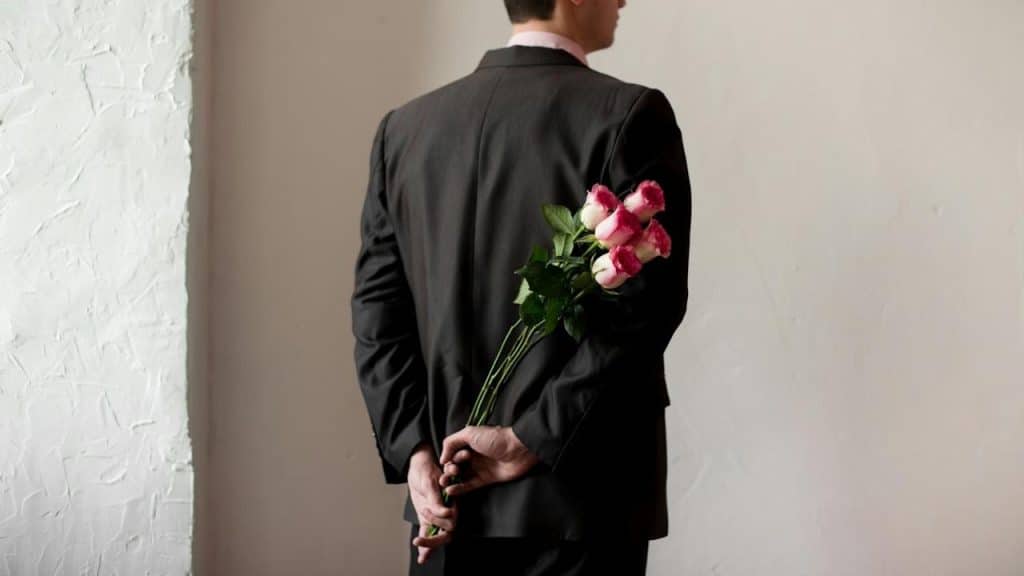 A man in a suit holding a bouquet of pink and white roses behind his back.