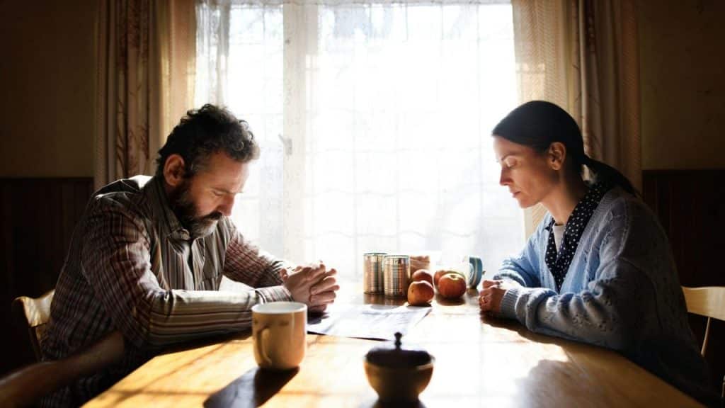 A man and woman sit at a table with heads bowed before a bright window.