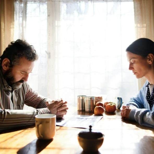 A man and woman sit at a table with heads bowed before a bright window.