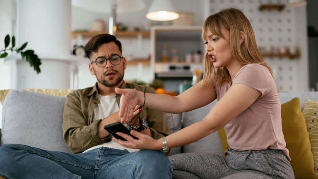 A woman speaks intensely while gesturing toward a smartphone held by a man on a sofa.