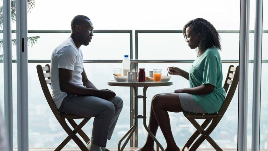 A man and woman sit at a small balcony table eating breakfast in silence.