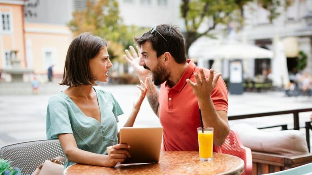 A man and woman argue intensely while sitting at an outdoor table with a tablet.