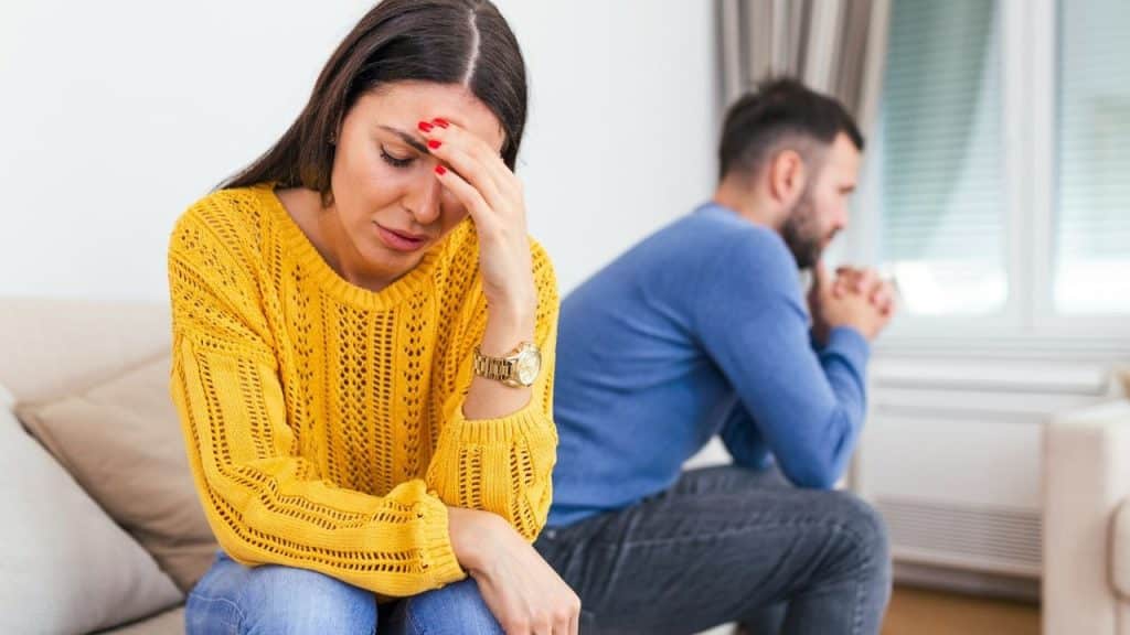 A woman in a yellow sweater looks down while a man sits blurred behind her.