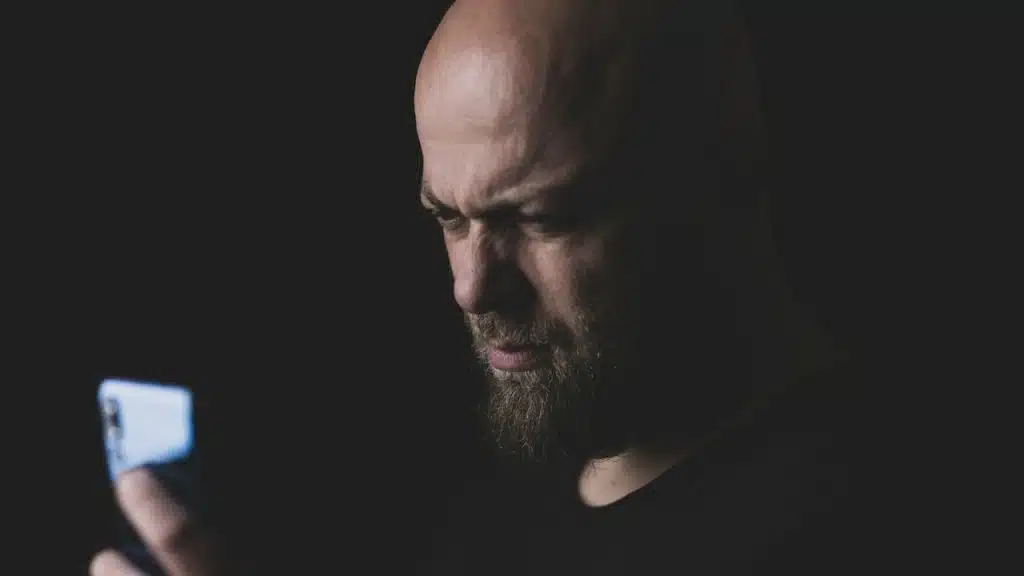 A bald man with a beard looks intensely at a glowing smartphone in a dark room.