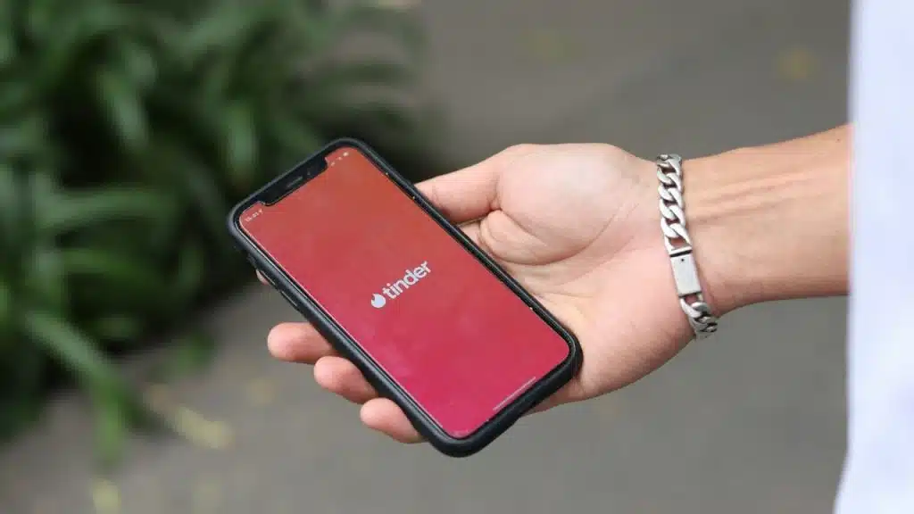 A hand holds a smartphone displaying the Tinder app logo against a blurred outdoor background.