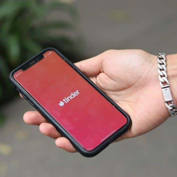 A hand holds a smartphone displaying the Tinder app logo against a blurred outdoor background.