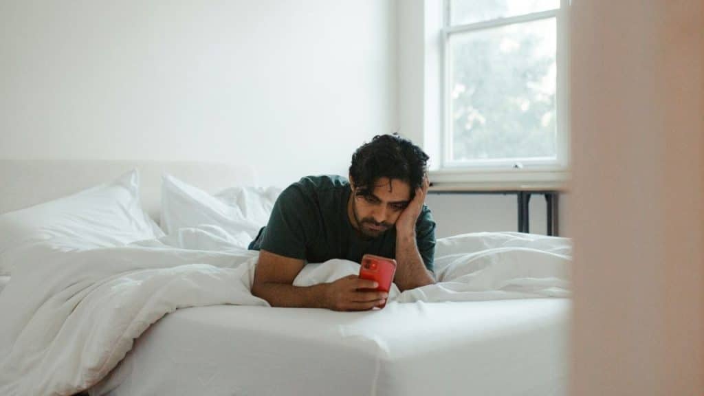 A man lies in bed and rests his head on his hand while looking at a smartphone.