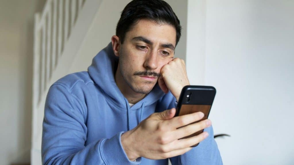 A man in a blue hoodie rests his head on his hand while looking at a phone.