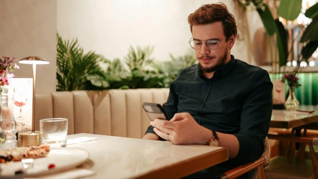 A man wearing glasses looks down at a smartphone while sitting at a restaurant table.
