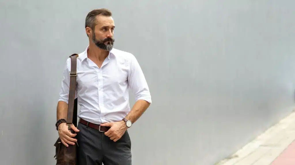 A man in a white shirt and gray trousers stands outside against a gray wall.