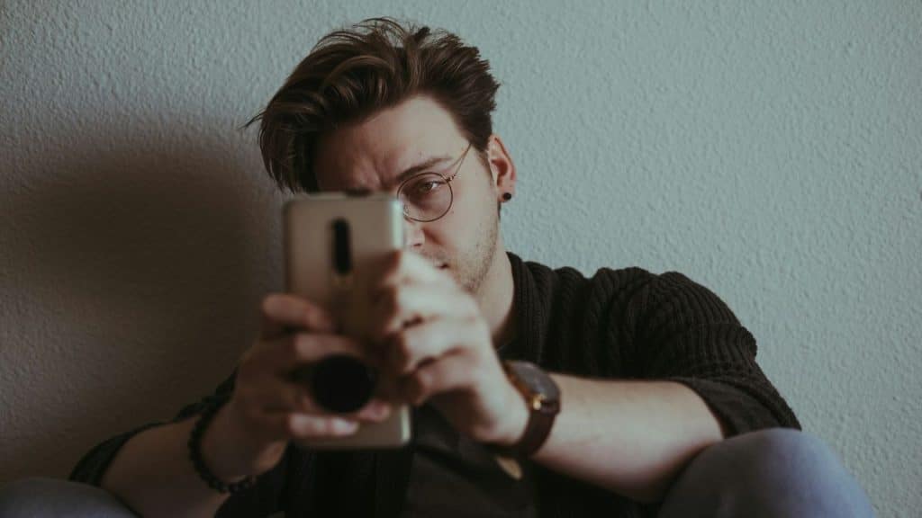 A man wearing glasses holds a smartphone up to his face while sitting against a wall.