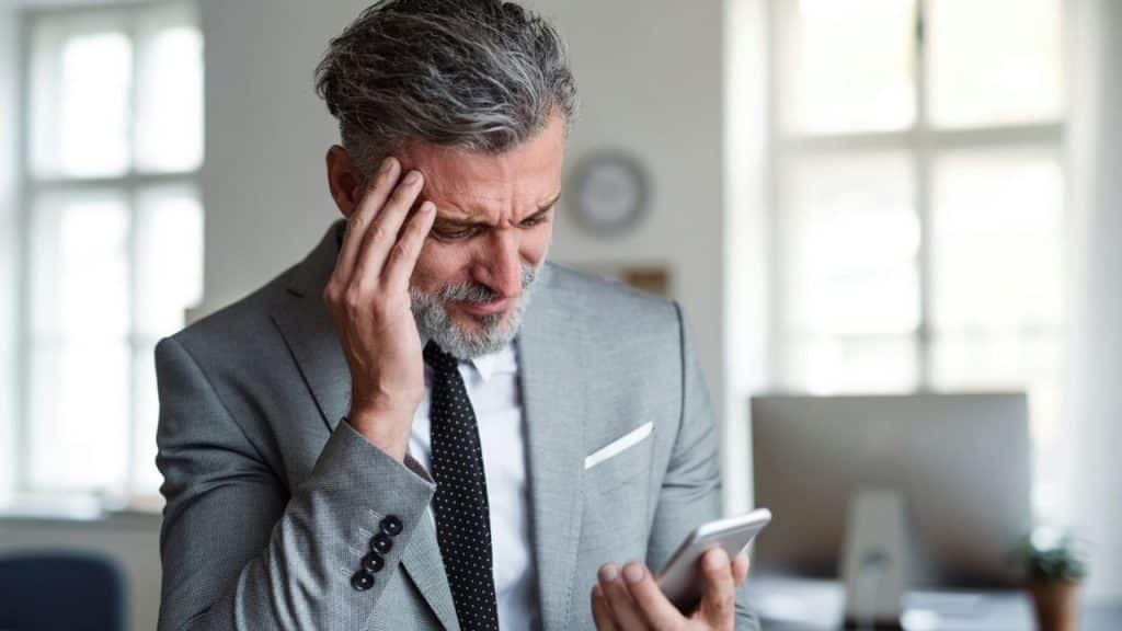 A man in a suit looks at a smartphone while holding his hand to his head.