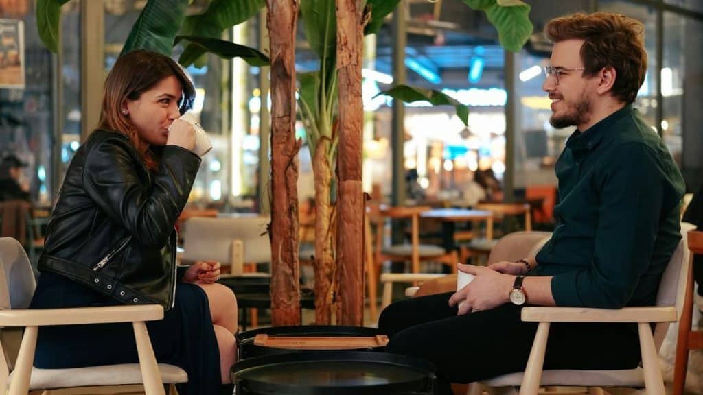 A man and woman sit in a cafe, talking and holding mugs across a table.
