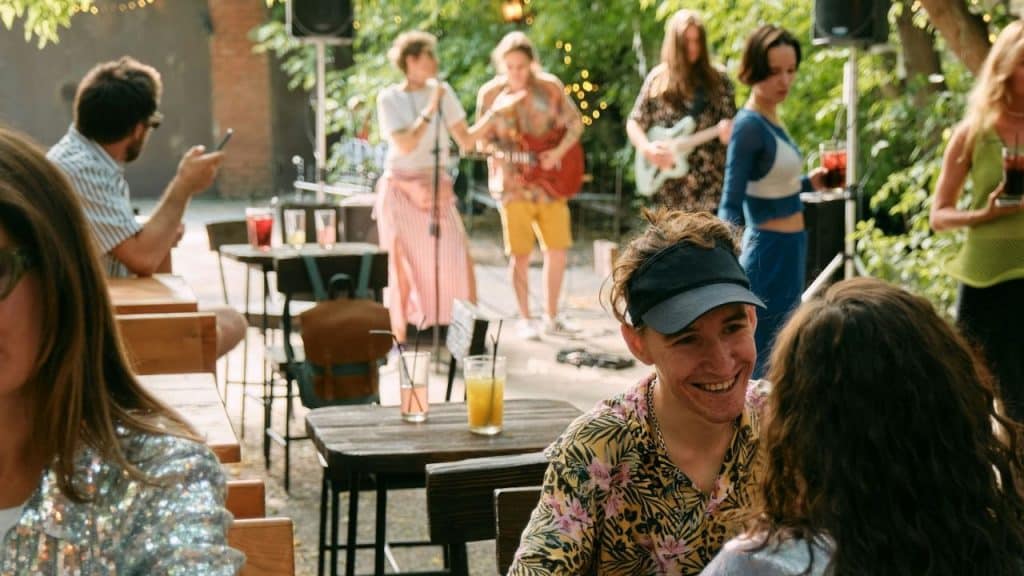 A smiling man in a floral shirt sits at an outdoor table with a band playing.