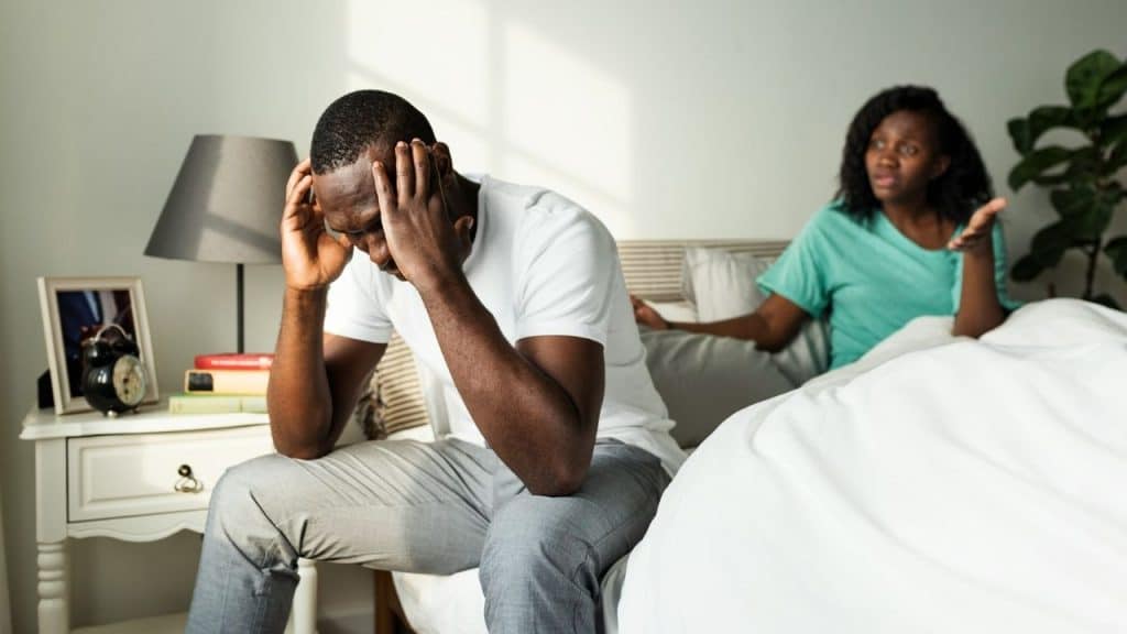 A man sits on a bed with his head in his hands while a woman talks.