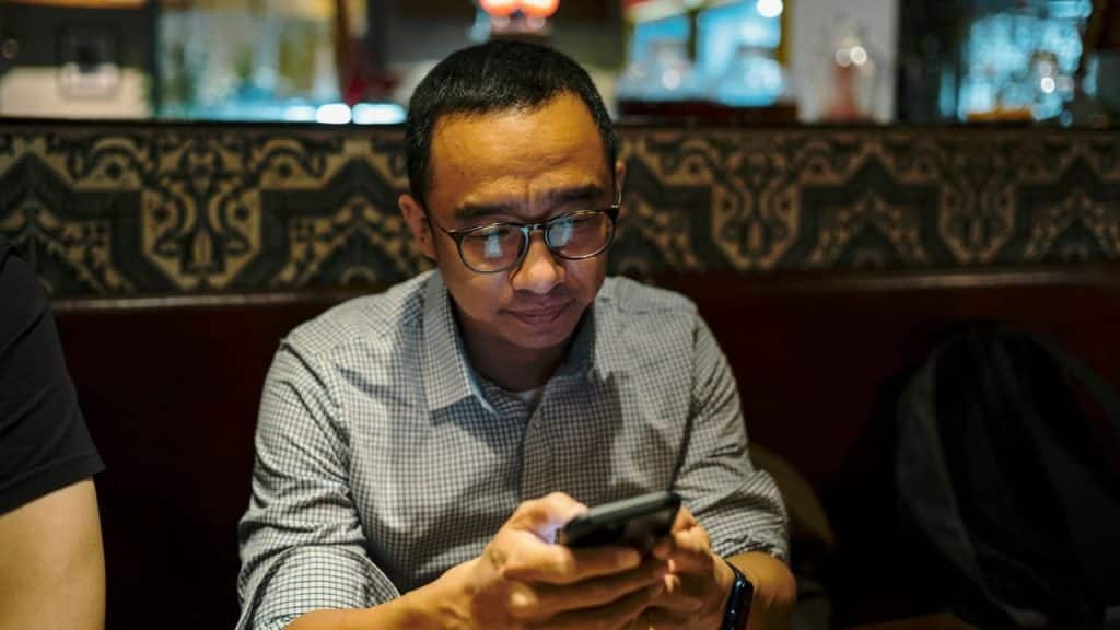 A man wearing glasses looks down at a glowing smartphone screen while sitting in a restaurant.