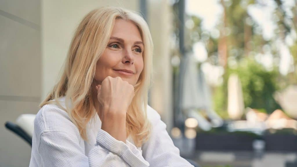 A blonde woman in a white robe rests her chin on her hand and smiles.