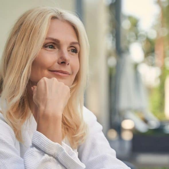 A blonde woman in a white robe rests her chin on her hand and smiles.