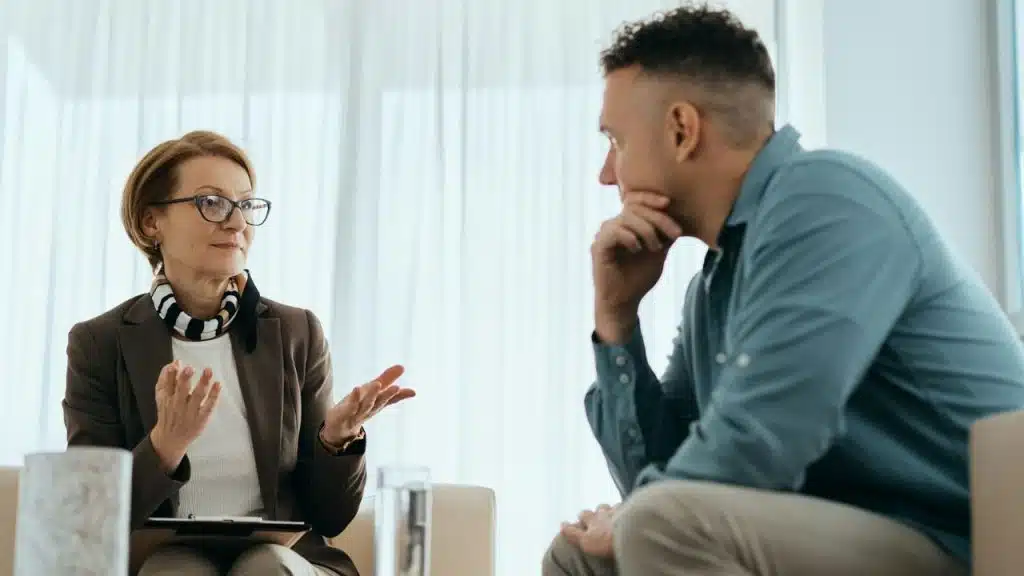 A woman with glasses and a brown blazer gestures while talking to a man.
