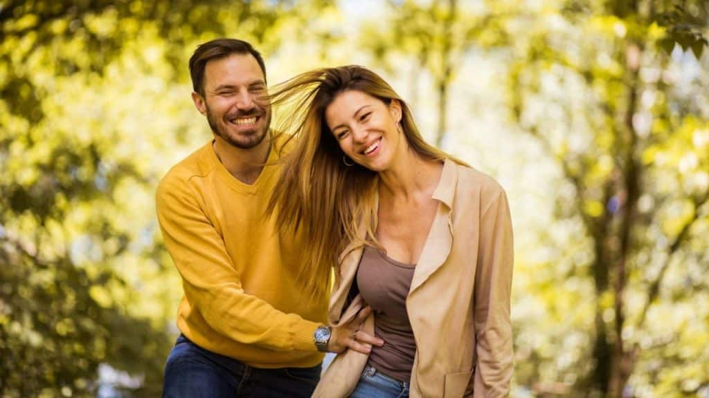 A man and woman laugh outdoors while the woman’s hair blows across the man’s face.