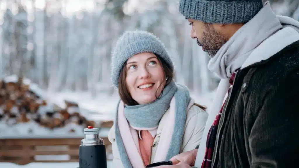 A woman and a man wearing winter clothes talking outdoors in a snowy forest.