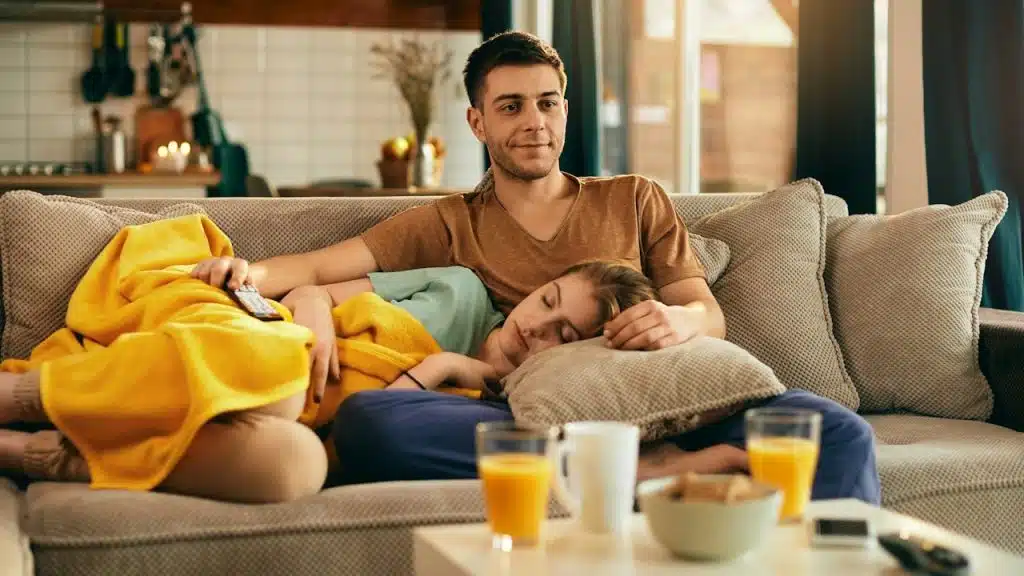 A woman sleeping with her head resting in a man’s lap as he watches TV while they occupy a couch.