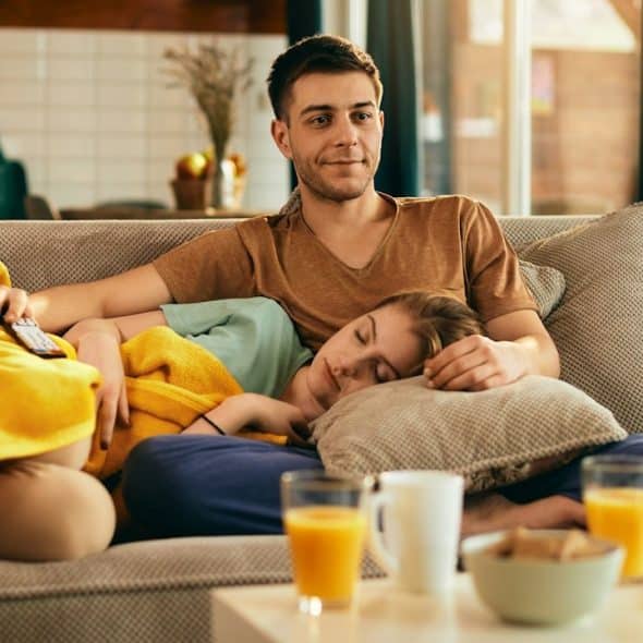 A woman sleeping with her head resting in a man’s lap as he watches TV while they occupy a couch.