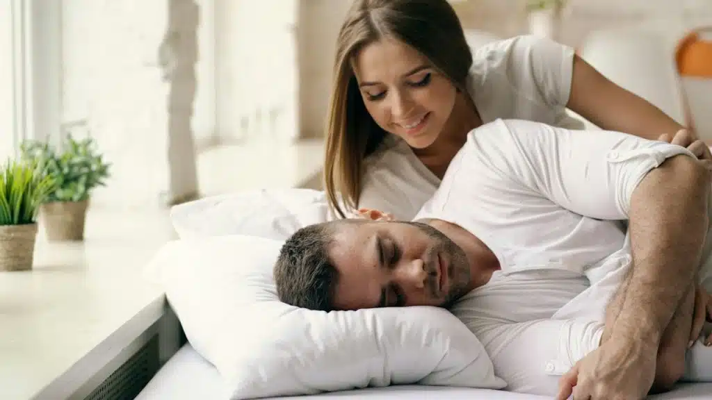 A woman and watching a man sleep while they lie on a bed.