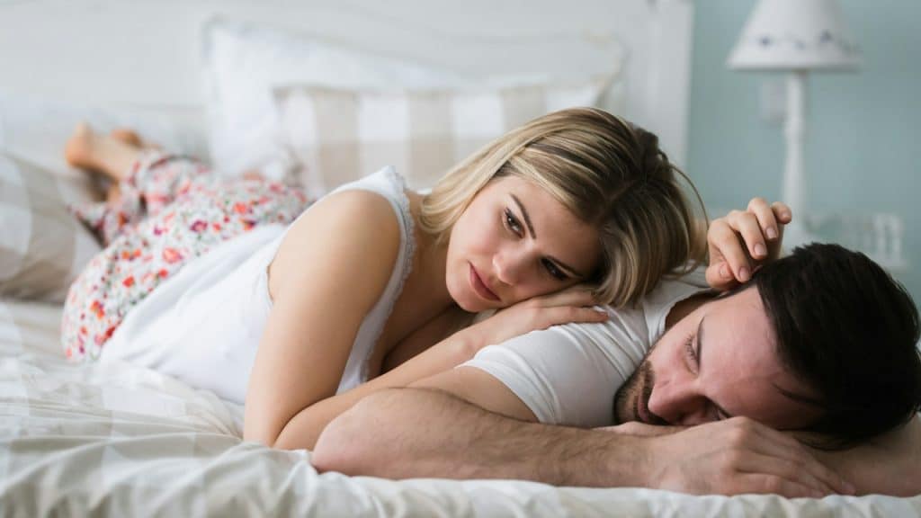 A thoughtful woman lying on a bed while her husband is sleeping next to her.