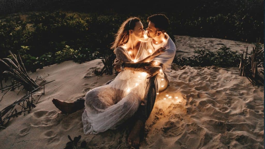 A man and woman looking at each other sitting on white sand during sunset.