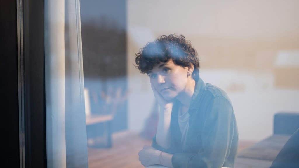 A person resting their head on a hand while gazing pensively through a window.