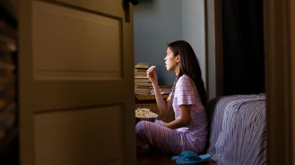 A woman in pajamas sitting on the floor, eating popcorn and watching television.
