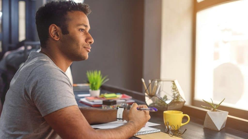 A man sitting at a desk by a window, holding a pen and looking thoughtfully outside.