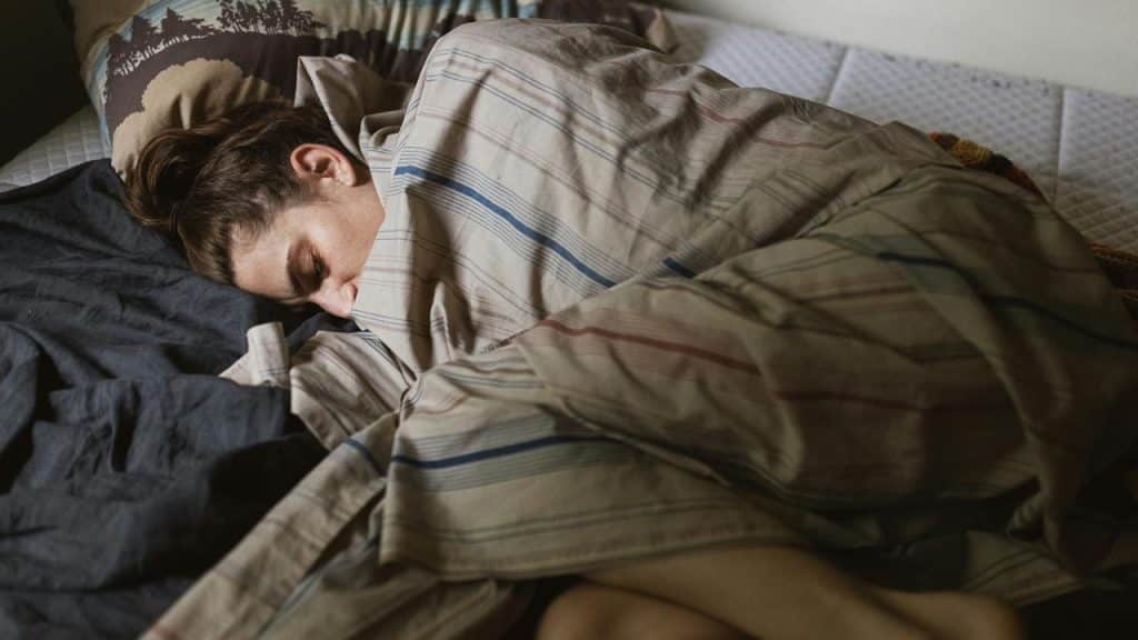 A person sleeping curled up under striped blankets on a bed.