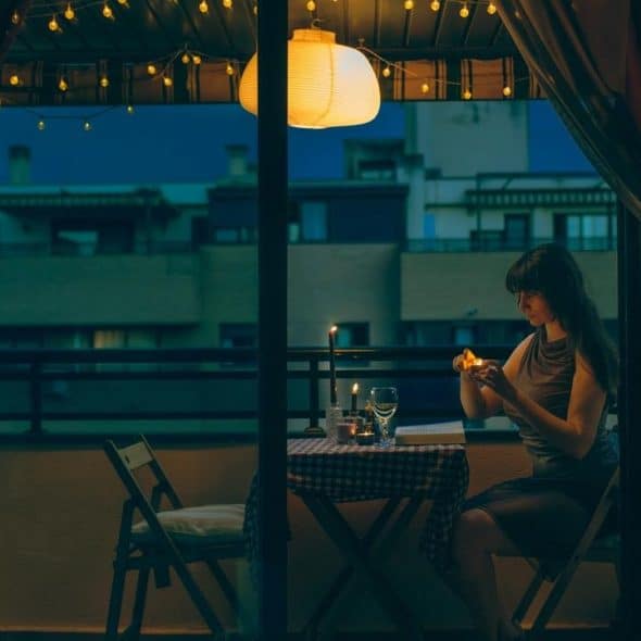 A person lighting a candle at a small balcony table under string lights at night.