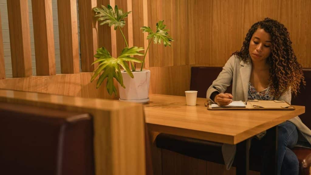 A woman writing in a notebook at a wooden table with a potted plant nearby.