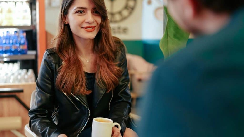 A woman smiling and holding a coffee mug while talking to someone in a café.