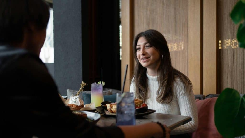 A woman smiling while chatting across a table with food and drinks in a café.