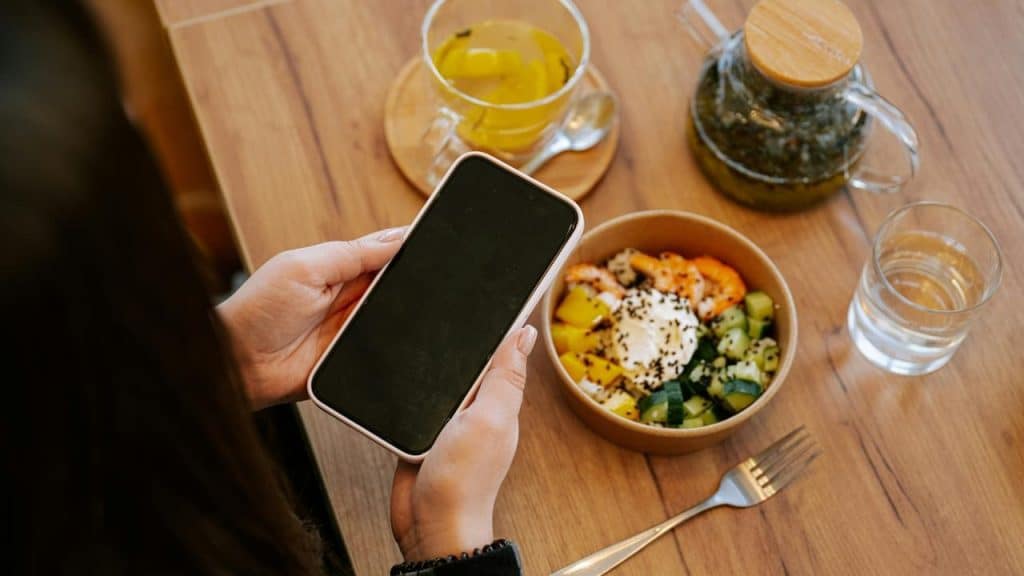 A person holding a smartphone above a bowl of colorful food on a wooden table.