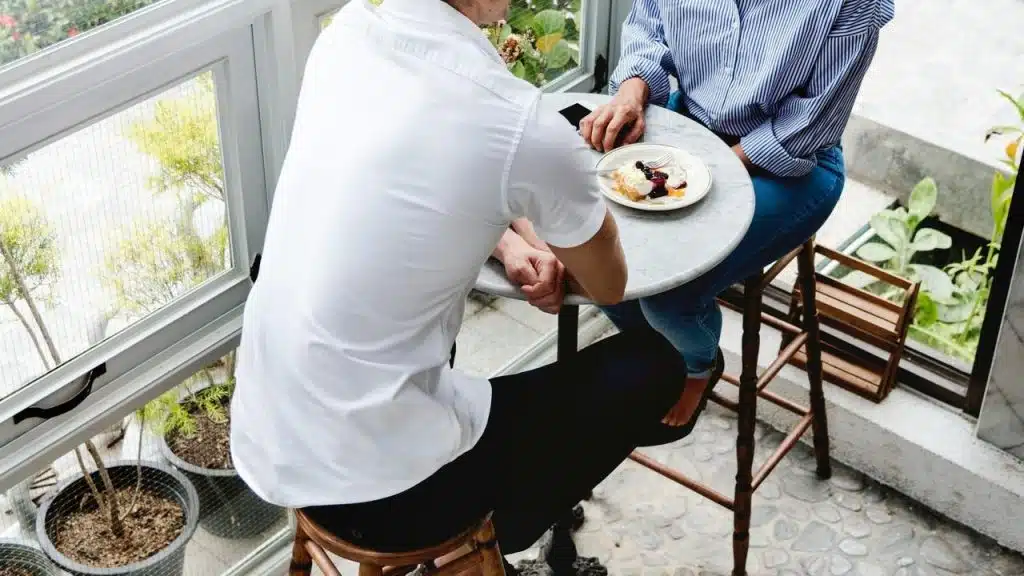 A couple sitting at a small round table by a window, sharing dessert.