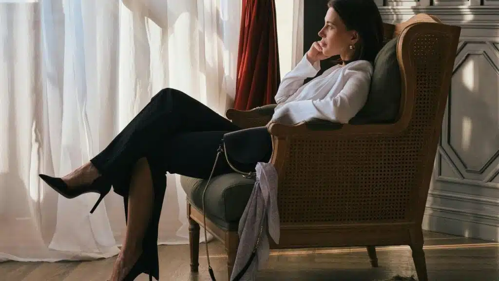A woman in a white blouse and black heels sitting thoughtfully in an armchair by a window.