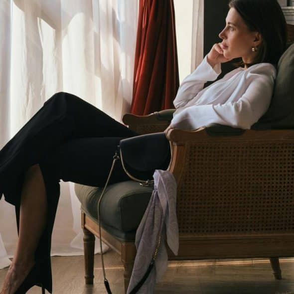 A woman in a white blouse and black heels sitting thoughtfully in an armchair by a window.