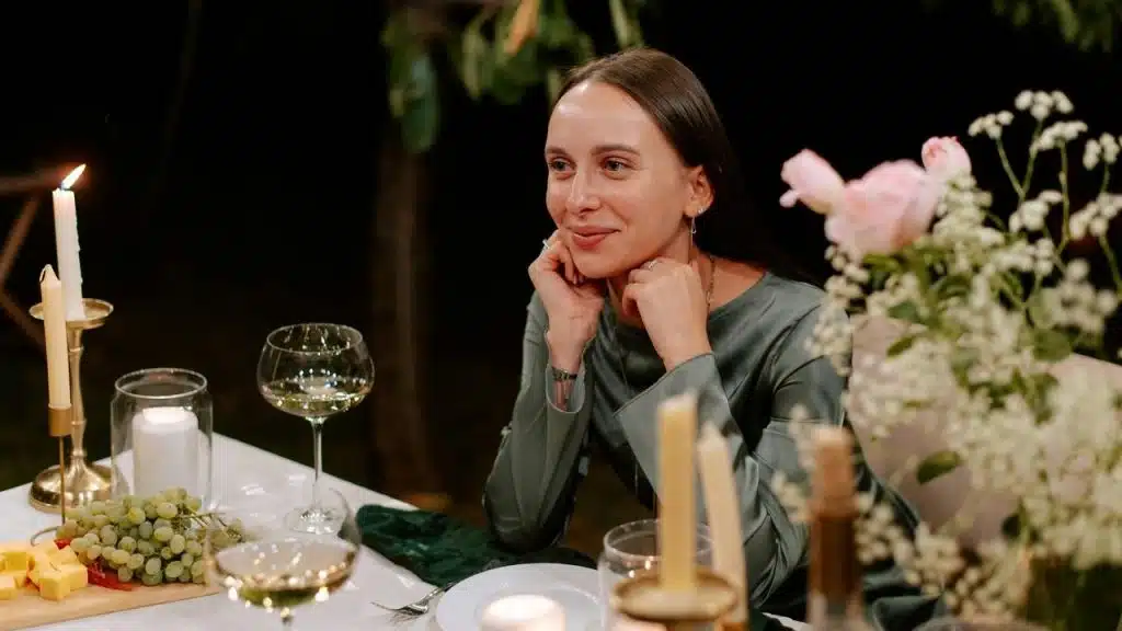 A woman smiling at a candlelit dinner table with wine, grapes, and flowers.